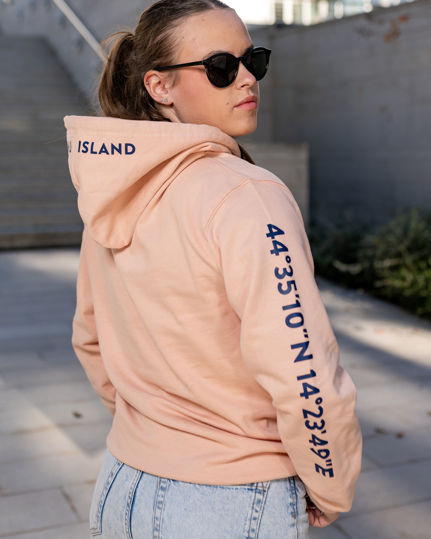 A girl wearing a peach hoodie with navy blue screen printed Lošinj island coordinates on the sleeve and the words Lošinj island on the hood, standing outdoors.