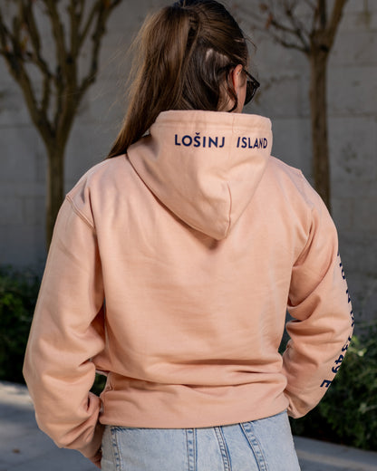 A girl wearing a peach hoodie with navy blue screen printed Lošinj island coordinates on the sleeve and the words Lošinj island on the hood, standing outdoors.