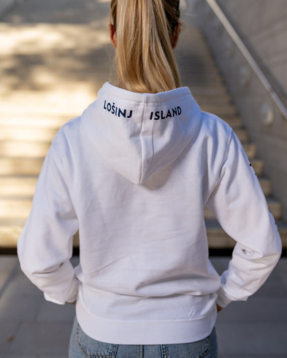 A girl wearing a blue hoodie with white screen printed Lošinj island coordinates on the sleeve and the words Lošinj island on the hood, standing outdoors. Original design by Bongolala.