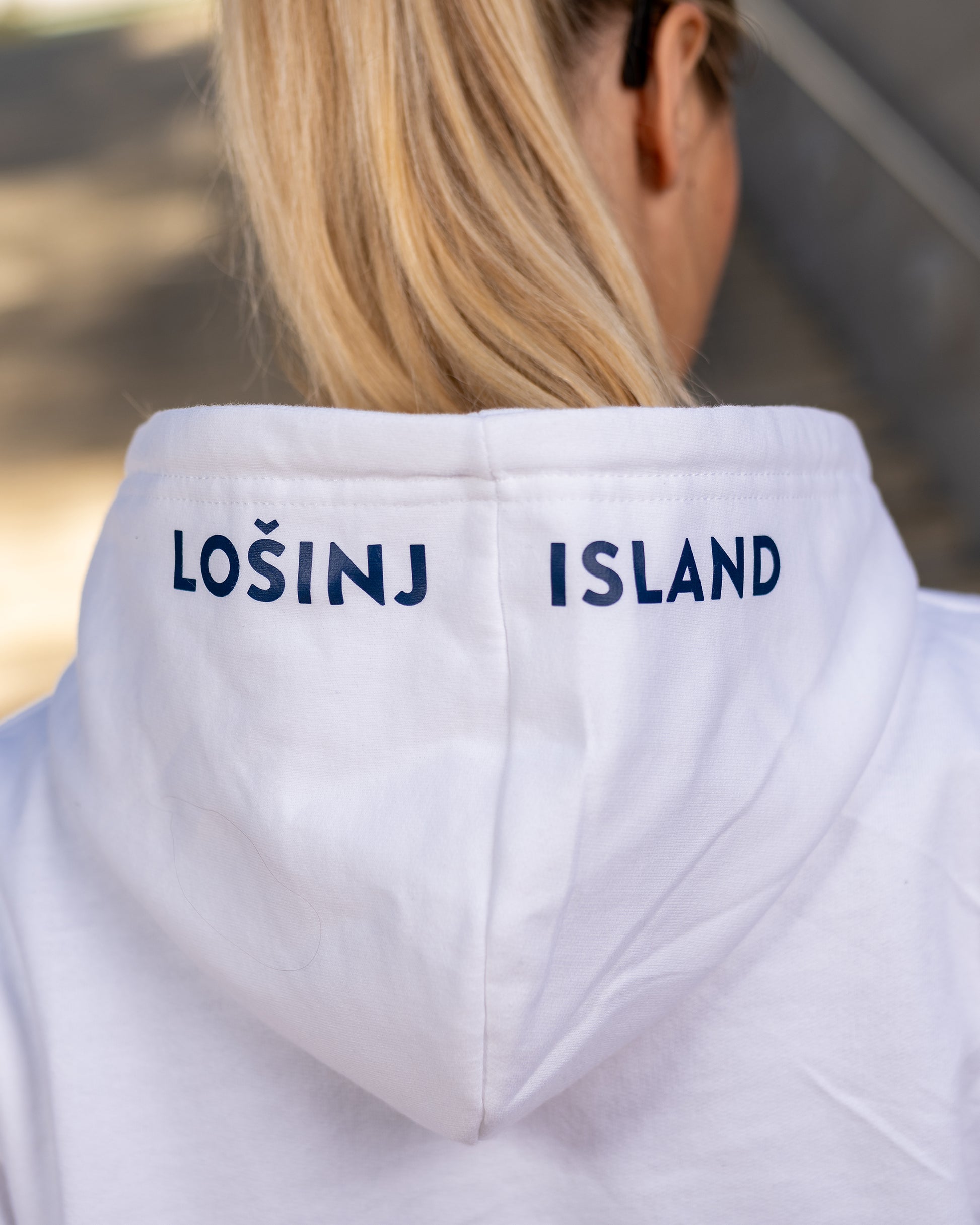 A girl wearing a blue hoodie with white screen printed Lošinj island coordinates on the sleeve and the words Lošinj island on the hood, standing outdoors. Original design by Bongolala.
