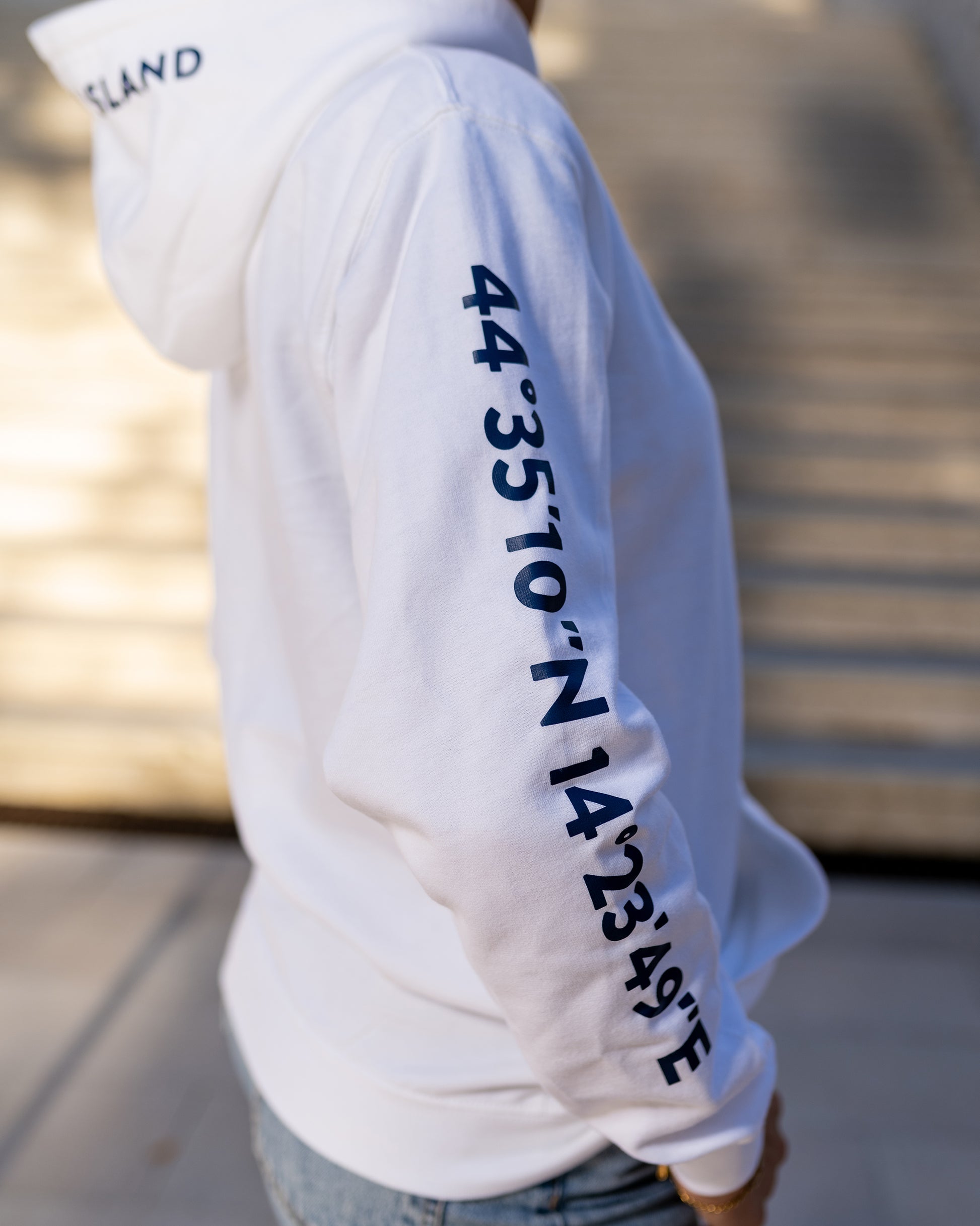 A girl wearing a blue hoodie with white screen printed Lošinj island coordinates on the sleeve and the words Lošinj island on the hood, standing outdoors. Original design by Bongolala.