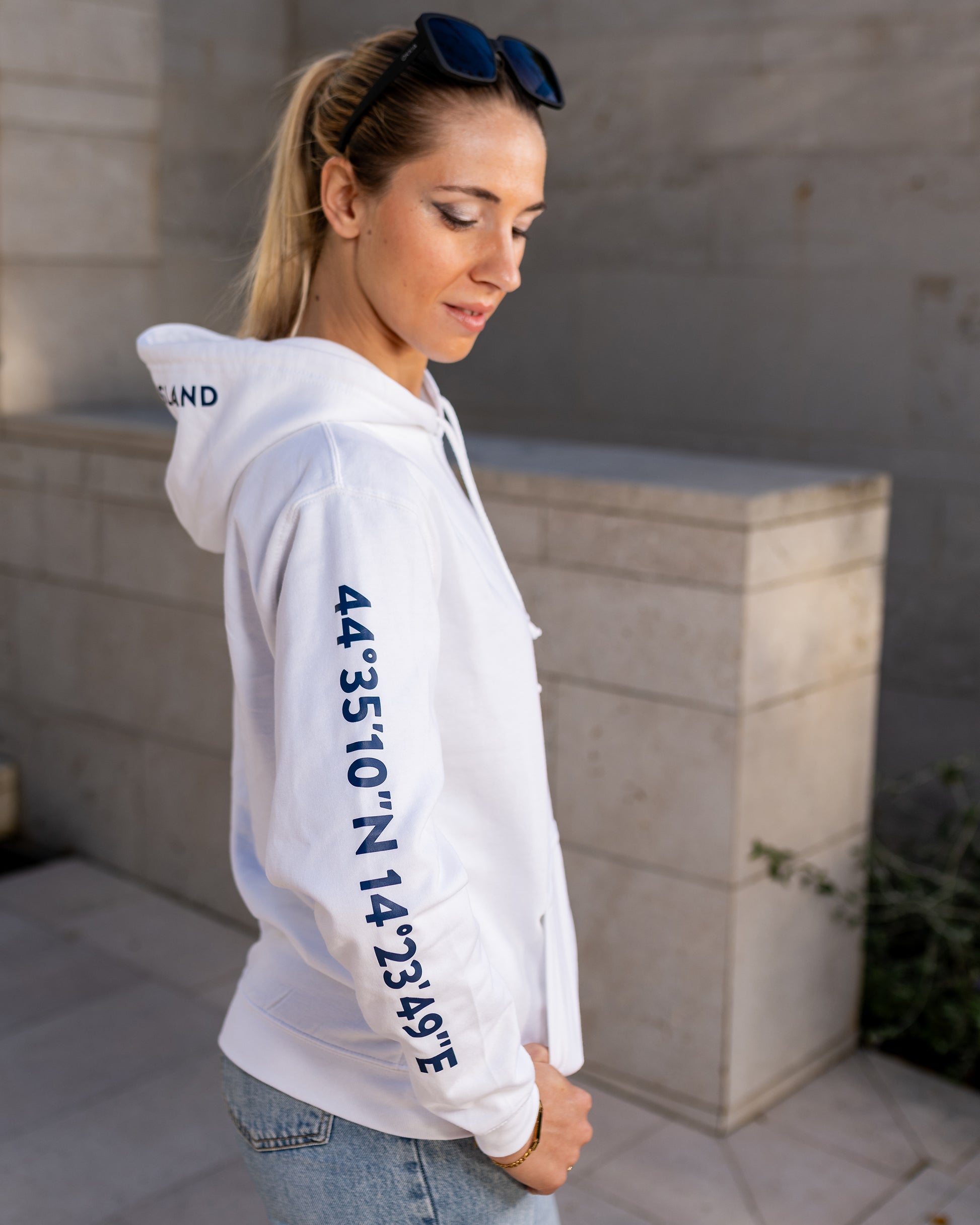 A girl wearing a blue hoodie with white screen printed Lošinj island coordinates on the sleeve and the words Lošinj island on the hood, standing outdoors. Original design by Bongolala.