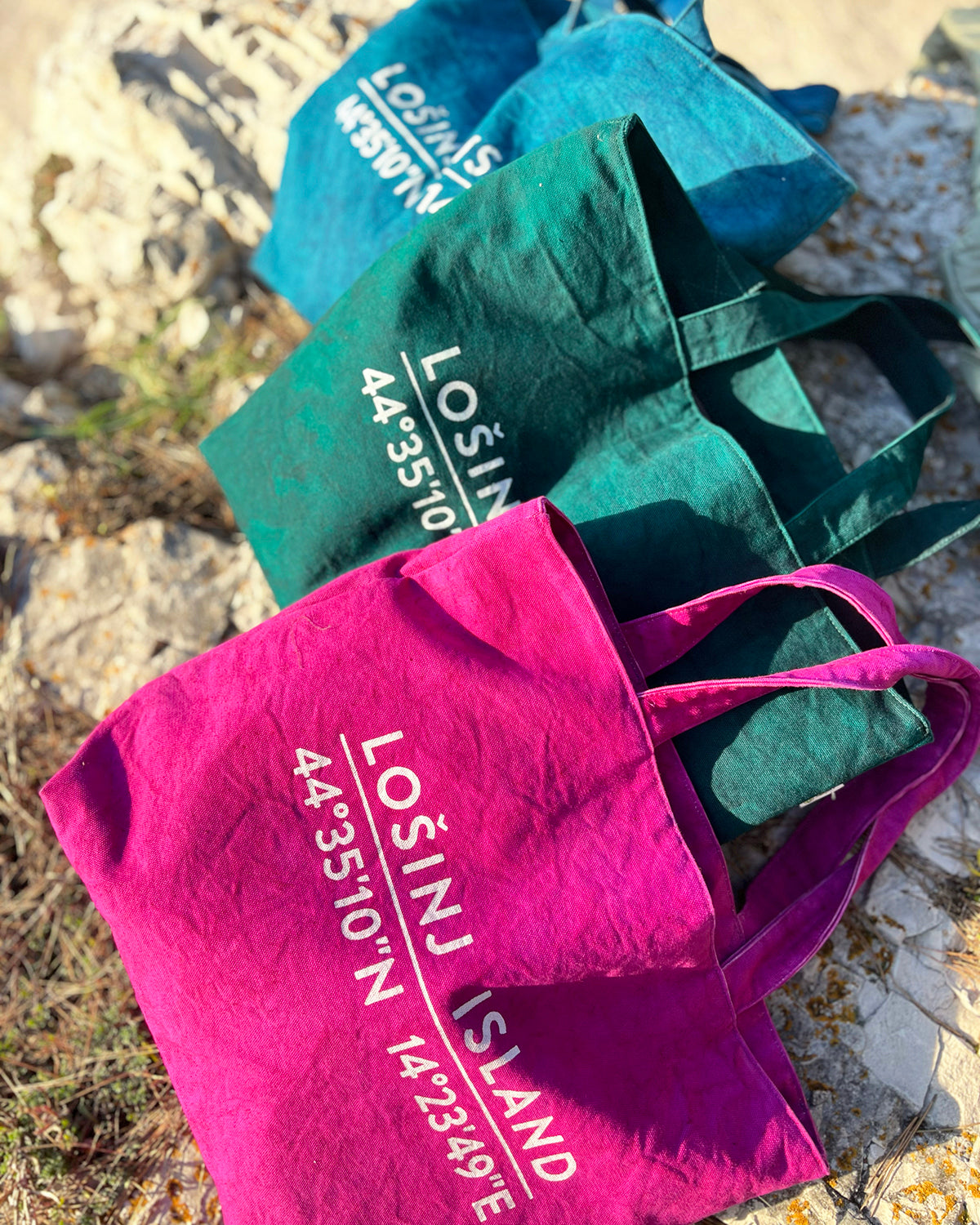Oversized cotton canvas shopper bags in pink, teal and botanical green with white screen printed Lošinj Island coordinates design on one side.
